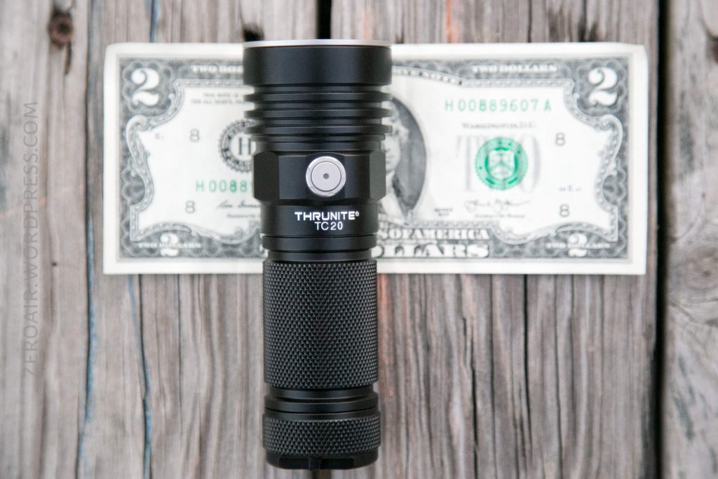 A black ThruNite TC20 flashlight is standing upright in front of a two-dollar bill on a weathered wooden surface. The flashlight is centered with its power button and branding visible.