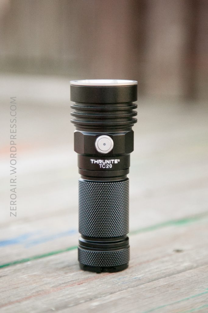 A black ThruNite TC20 flashlight with a textured grip and a silver button stands upright on a wooden surface. The background is out of focus.