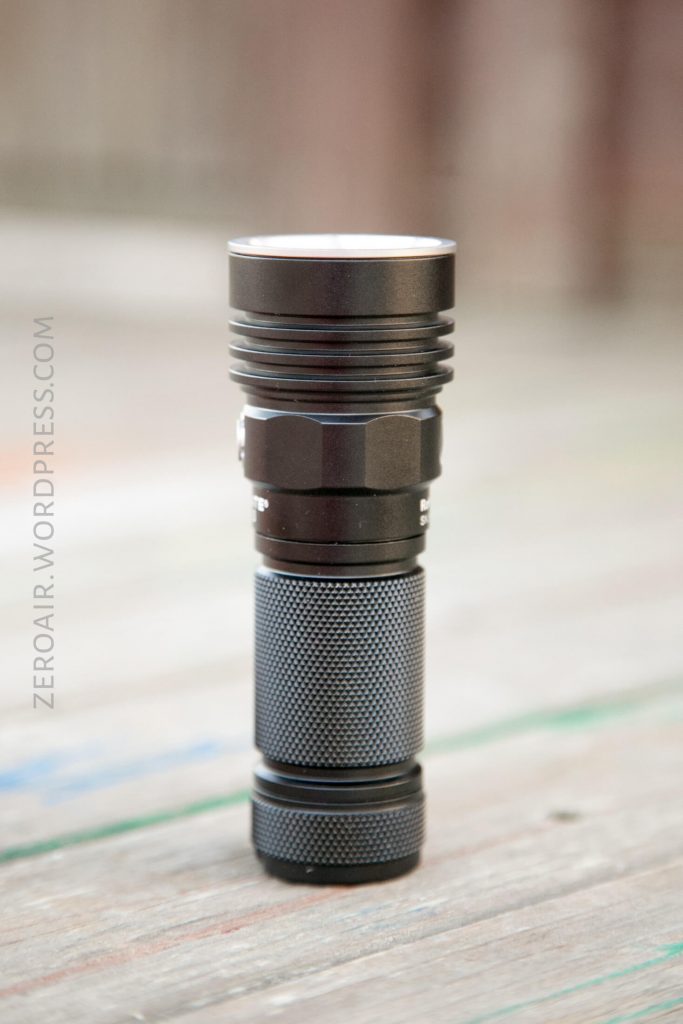 A black flashlight with a textured grip stands upright on a wooden surface. The background is out of focus, giving emphasis to the flashlight. The text ZEROAIR.WORDPRESS.COM is visible along the left side.