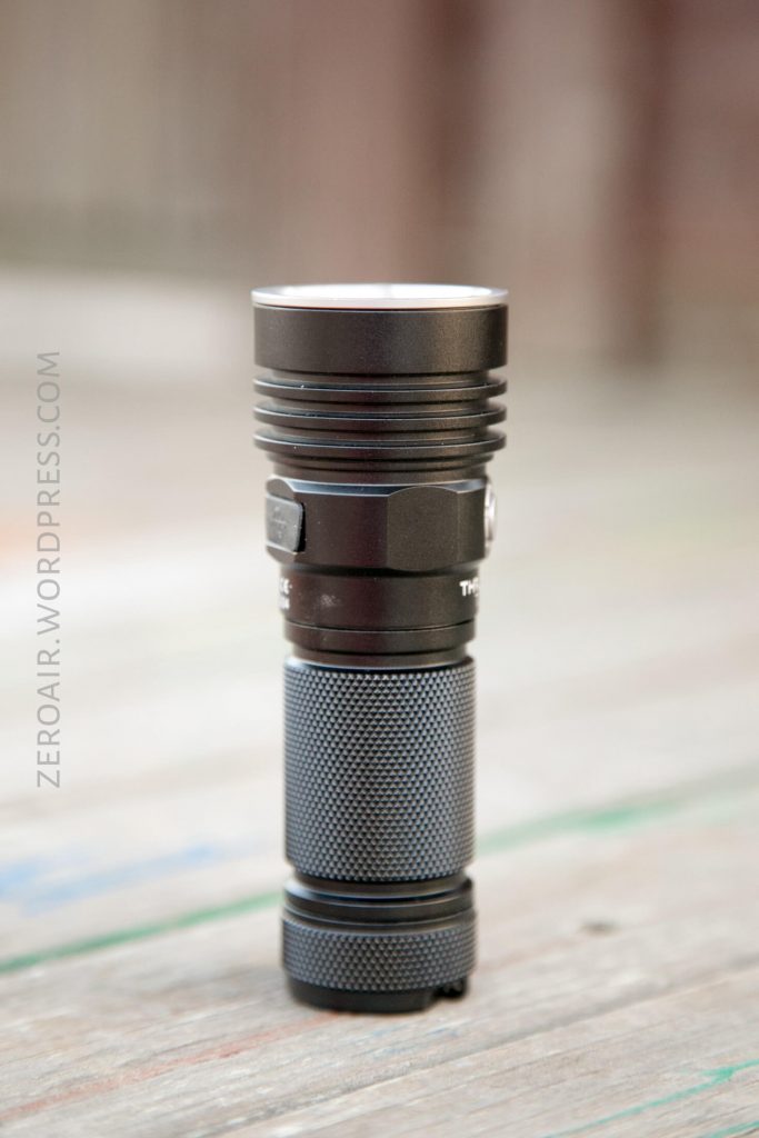A black, cylindrical LED flashlight with a textured grip is standing upright on a wooden surface. The background is blurred. The flashlight appears to be made of metal.