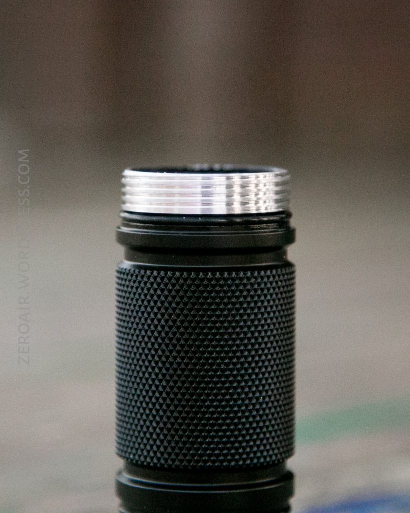 A close-up of a black, textured cylindrical object with visible silver threads at the top, possibly the body of a flashlight or similar device, standing upright on a blurred surface.