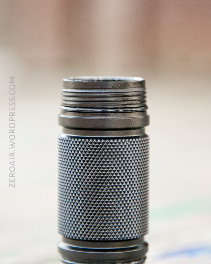 A close-up of a knurled metal flashlight body with visible threading at the top, standing upright on a blurred surface. The background appears out of focus, and text reads “zeroair.wordpress.com.”