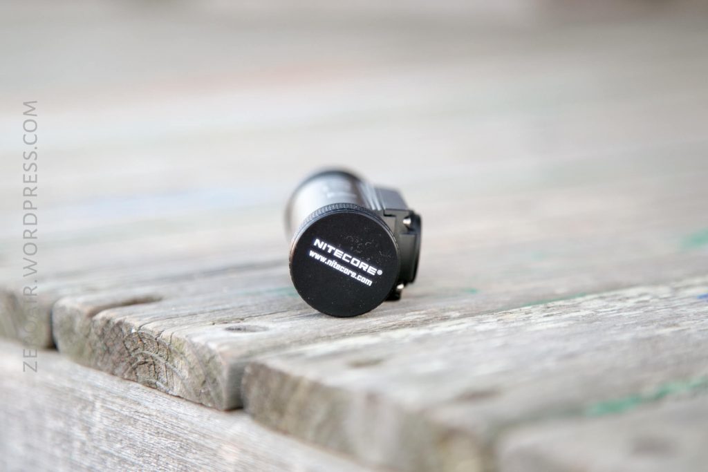 A small black cylindrical object with NITECORE and TM06S written on the end, resting on a weathered wooden surface. The background is blurred.