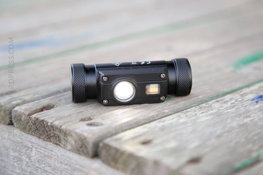 A black flashlight with two cylindrical ends and a central light is resting on a weathered wooden surface. The flashlight is turned on, emitting a white beam.