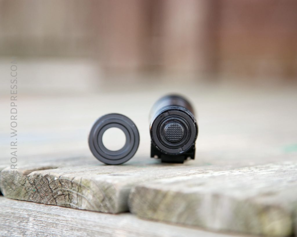 A close-up of a cylindrical black object with a textured button facing the camera, next to a detached circular cap, both resting on a weathered wooden surface.