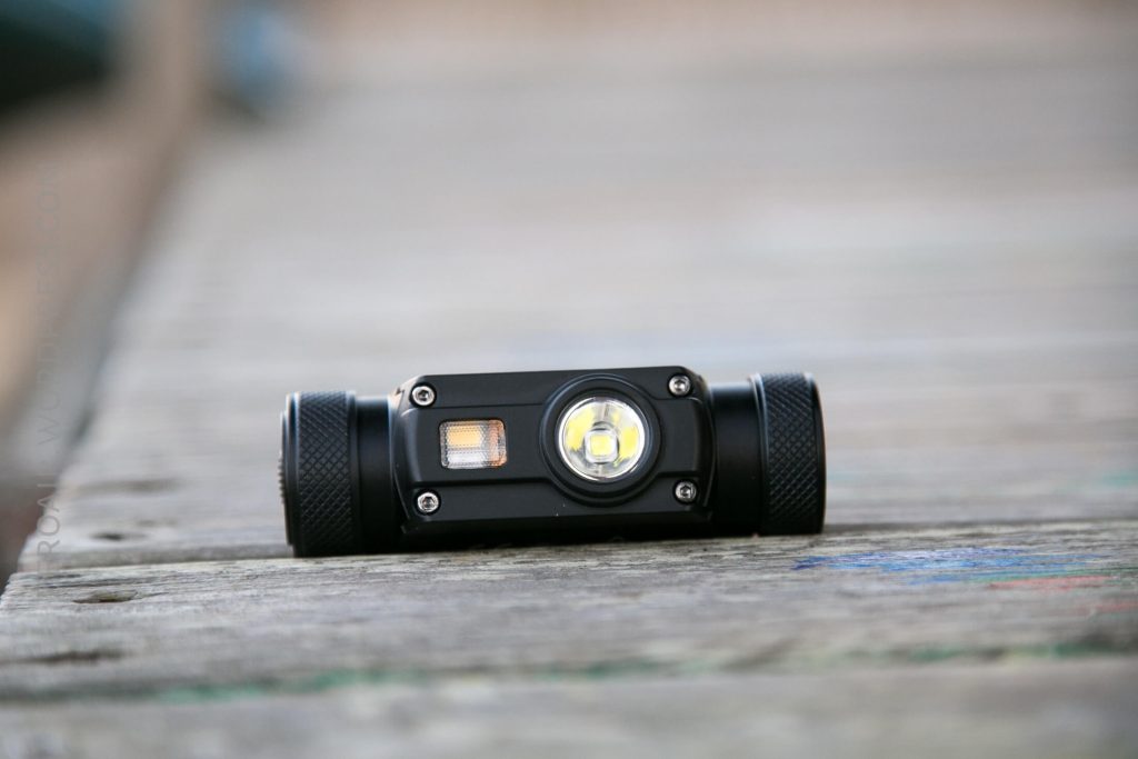A black handheld flashlight with a central LED bulb and a side panel rests on a wooden surface. The background is blurred, focusing attention on the flashlight.