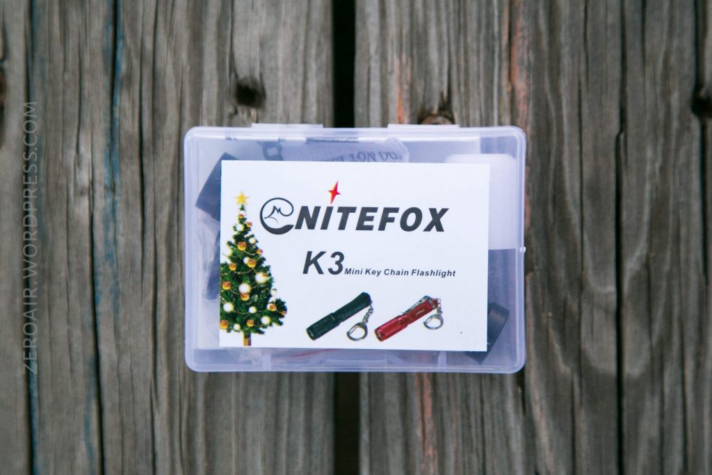 A clear plastic box with a label reading NITEFOX K3 Mini Key Chain Flashlight, featuring images of two flashlights, a Christmas tree, and a logo, placed on a wooden surface.