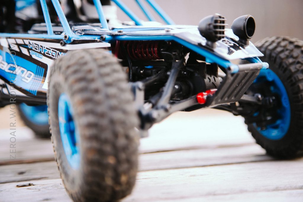 Close-up of the front suspension and wheels of a blue and black remote-controlled off-road vehicle, positioned on a wooden surface. The image focuses on the vehicle’s detailed parts and rugged tires.