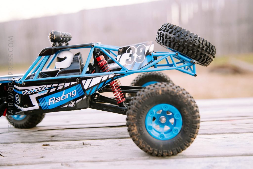 A blue and black remote-controlled off-road racing car with large, rugged tires is parked on a wooden surface outdoors, with a wooden fence in the background.