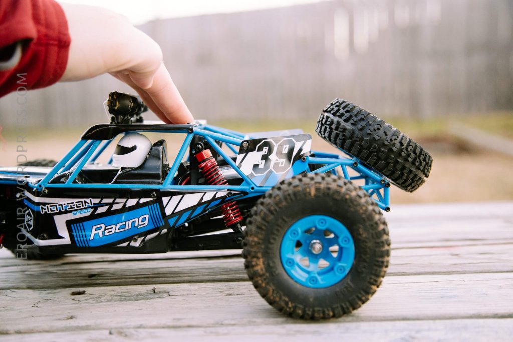 A person’s hand holds a blue and black remote-controlled racing car with the number 39 on the side, placed on a wooden surface outdoors with a blurred fence in the background.