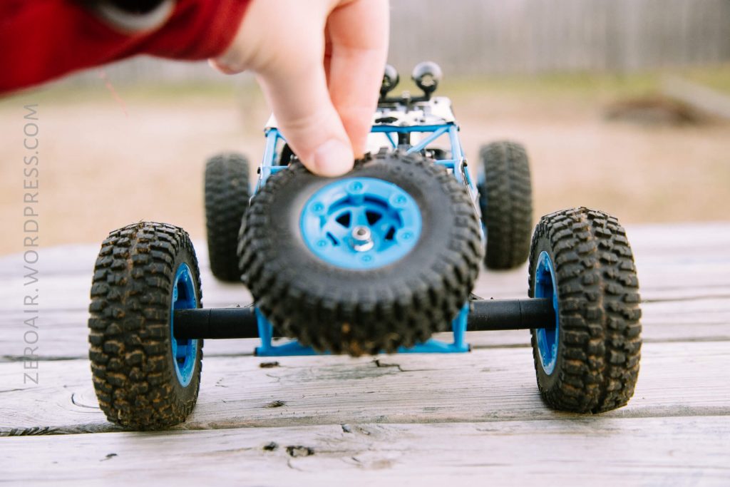 A hand places a spare tire onto the front of a blue remote control car with large, knobby tires, sitting on a wooden surface outdoors. The background is blurred.