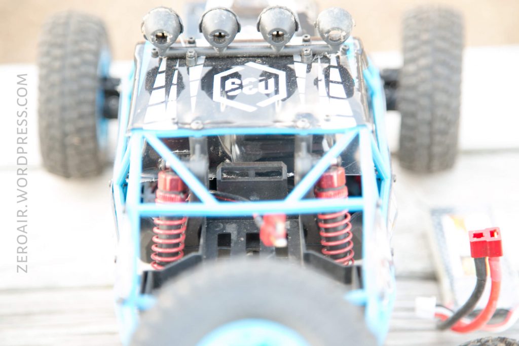 A close-up view of an RC car with blue frame, red coil shocks, and a black and white decal on the hood. Four spotlights are mounted on top. The image is partially out of focus. A connector wire is on the right side.
