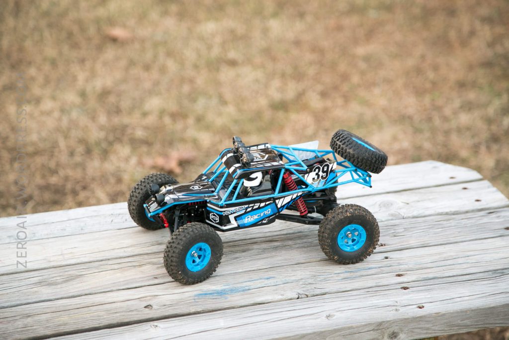 A blue and black remote-controlled off-road car with large tires and a spare tire on the back is placed on a weathered wooden surface, with dry grass in the background.