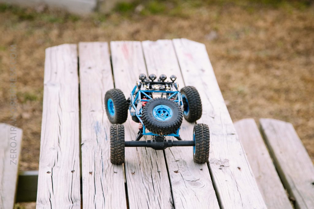 A blue remote-controlled off-road vehicle with large tires and a spare mounted on the hood is parked on a weathered wooden bench outdoors. The background shows dry grass.