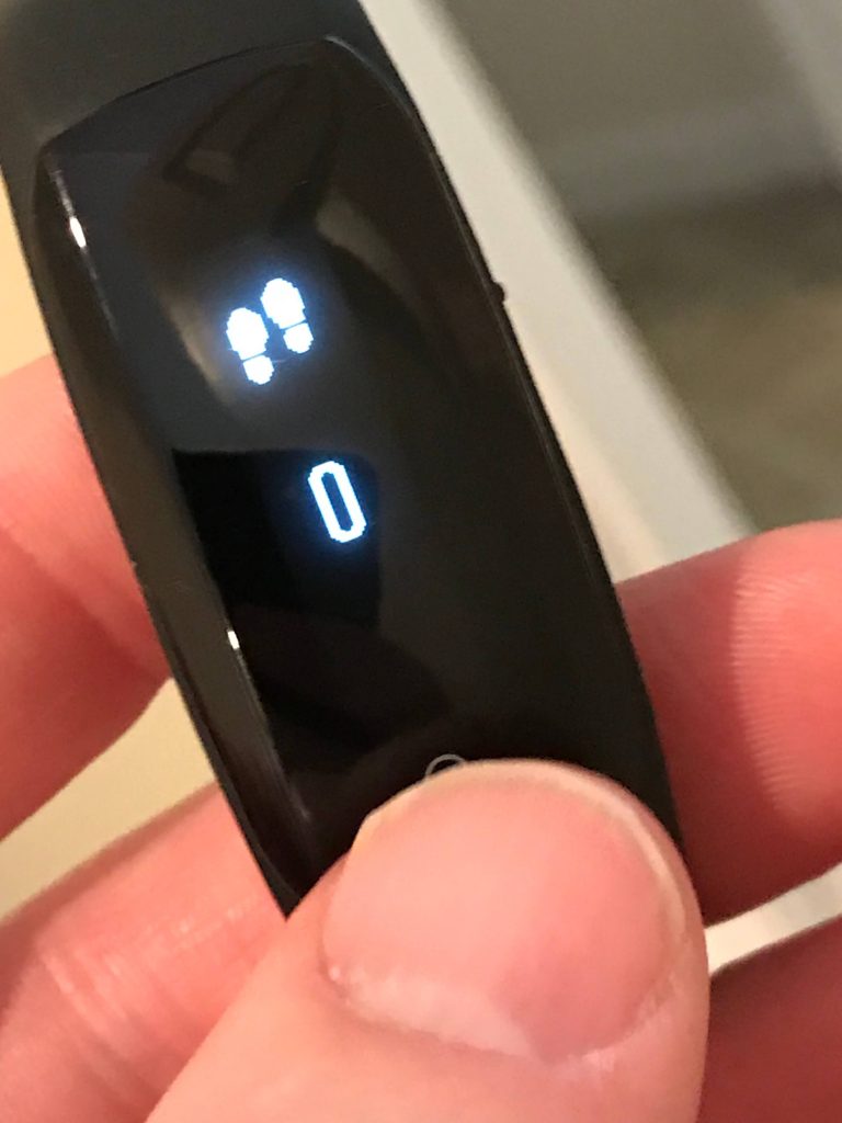 A close-up of a persons hand holding a black fitness tracker. The screen displays an icon of two shoes above the number zero, indicating no steps have been recorded.