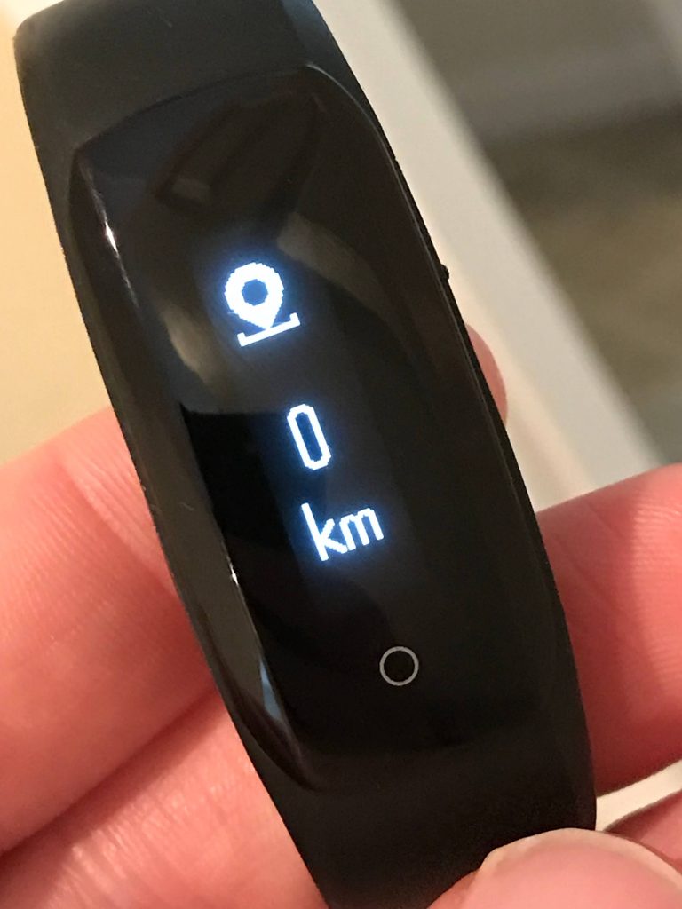 A close-up of a person holding a black fitness tracker. The tracker’s screen displays an icon, the number 0, and the unit km, indicating zero kilometers recorded.