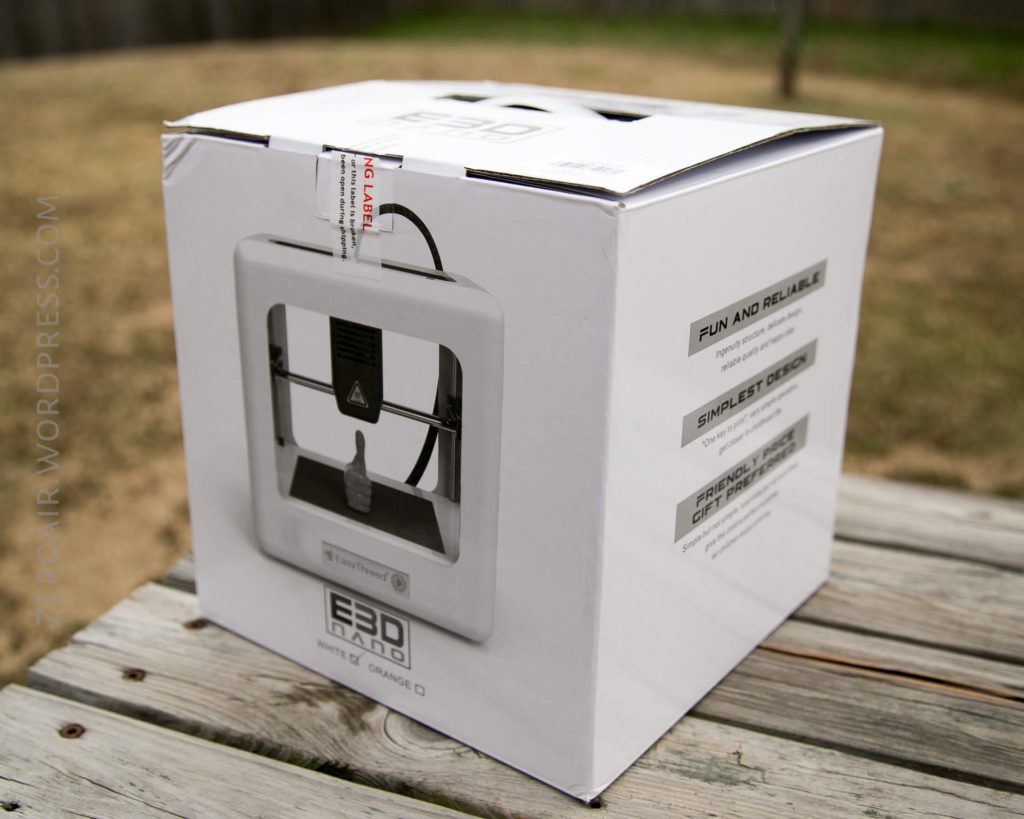 A white cardboard box for a 3D printer labeled “Micro+,” sitting on a wooden surface outdoors. The box displays an image of the 3D printer and product information on the side.