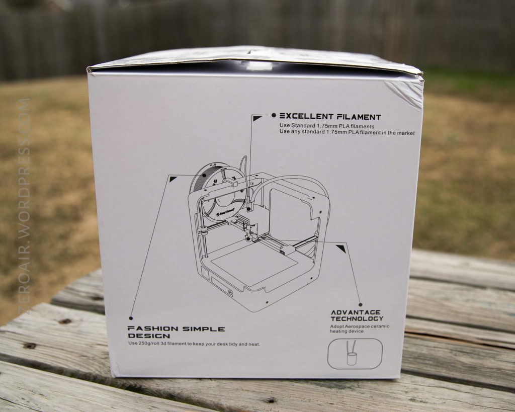 A white box with a line drawing of a 3D printer on it sits on an outdoor wooden surface. The box text highlights features such as filament compatibility and simple design. The background is a blurred grassy yard.