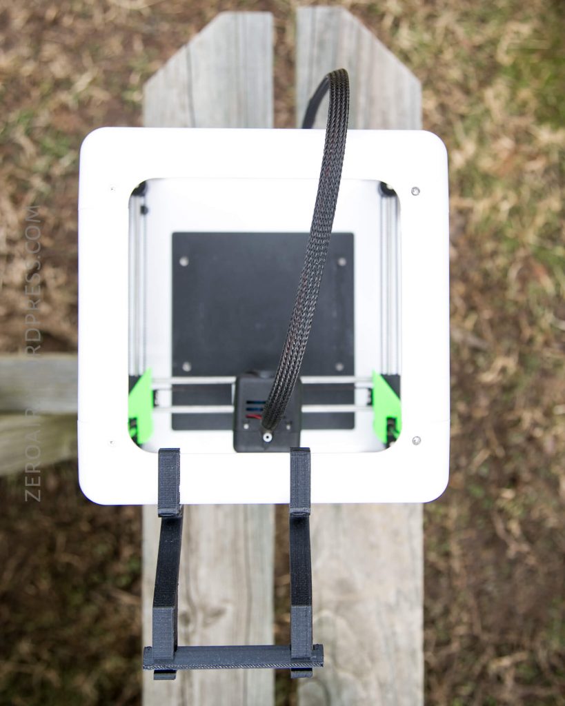 A top-down view of a white 3D printer with a black cable, sitting on a wooden outdoor surface with grass visible in the background.