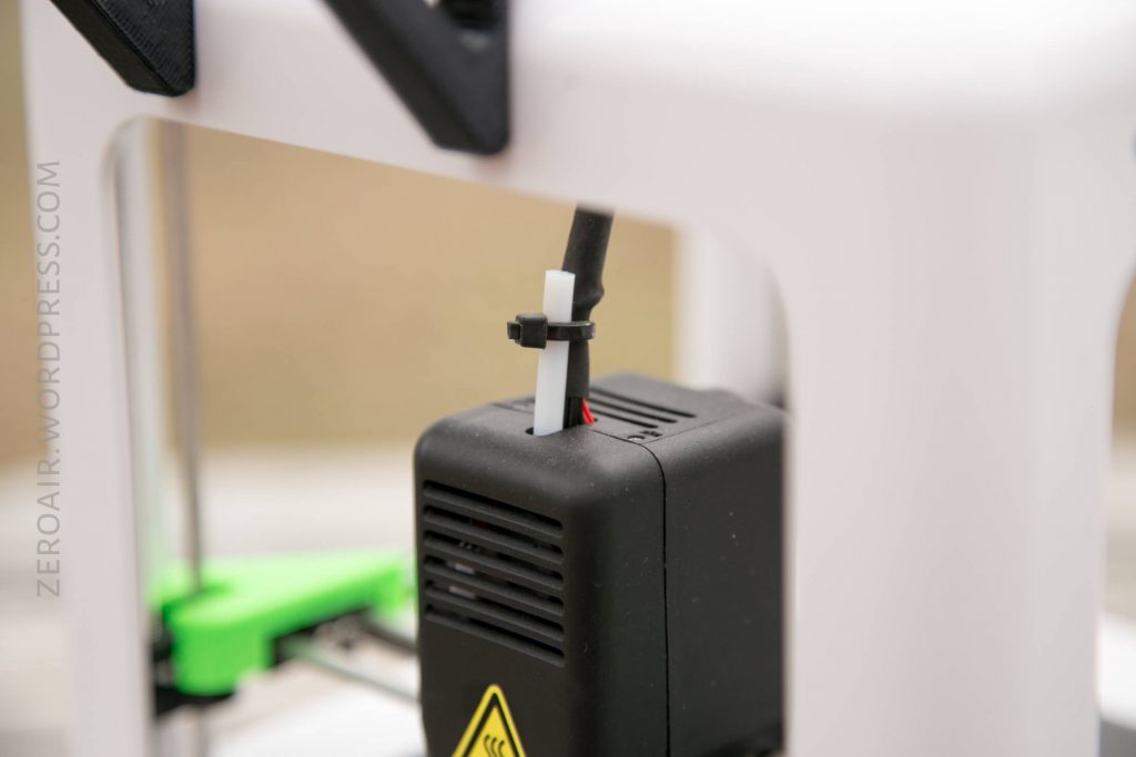 A close-up of a black component on a 3D printer, with a cable secured by a zip tie and a warning label partially visible on the front. The background is blurred.