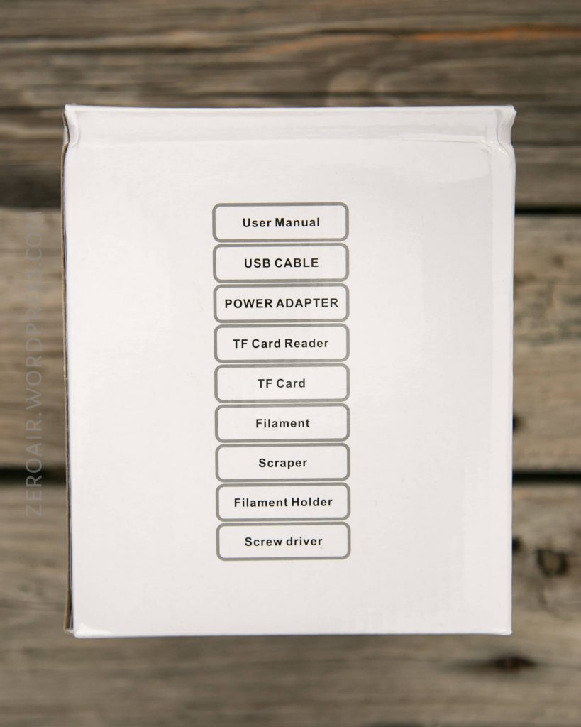 A white box with a printed list of contents: user manual, USB cable, power adapter, TF card reader, TF card, filament, scraper, filament holder, and screw driver. The box is placed on a wooden surface.