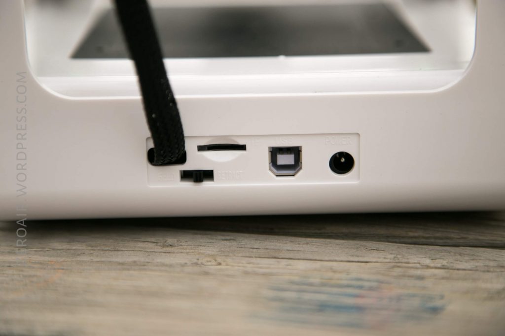 Close-up of the back of a white electronic device showing an SD card slot, a power switch, a USB-B port, and a round power input port, with part of a cable visible above the ports.