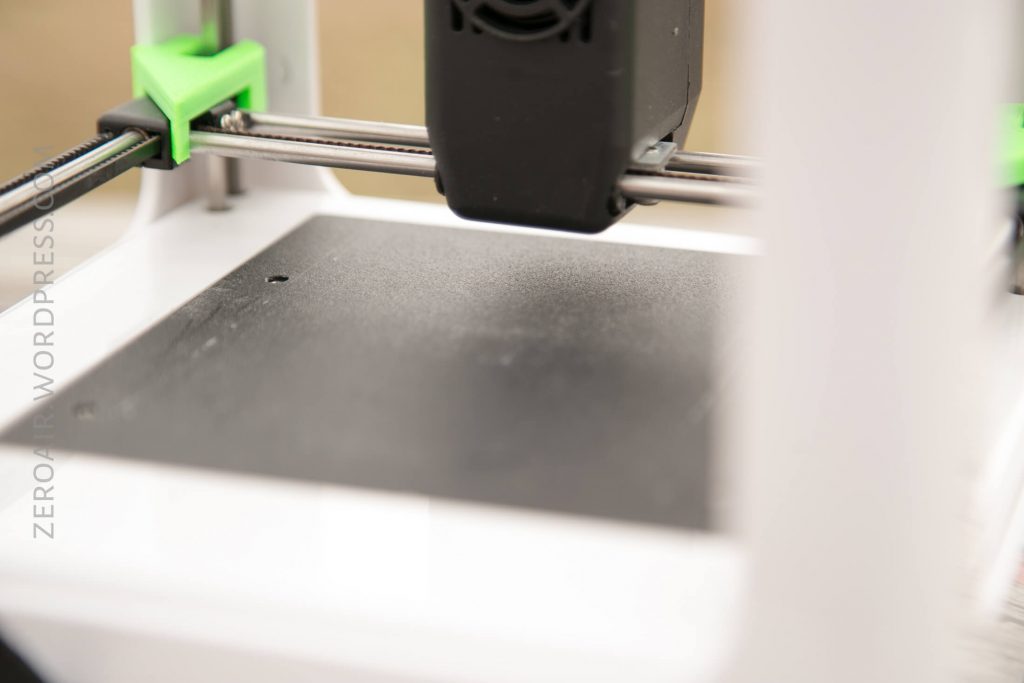 Close-up view of a 3D printer, focusing on the print bed and the print head above it, with green and black components visible. The machine appears to be idle.