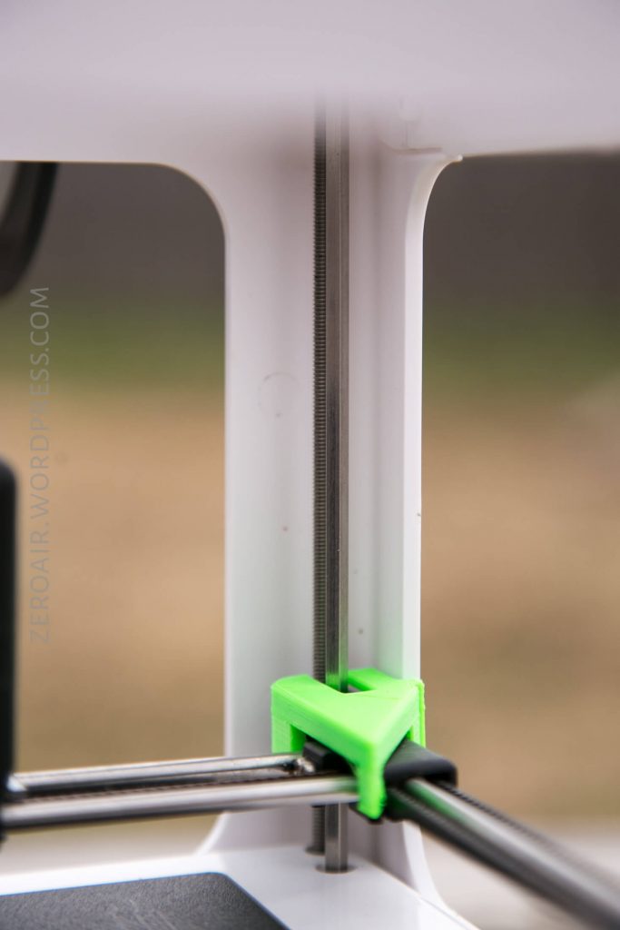 A close-up view of a 3D printer’s threaded rod and metal rail, with a green plastic component securing the rod in place. The background is blurred and part of a white frame is visible.