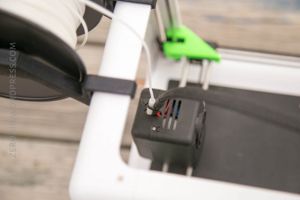 Close-up of the filament feeder mechanism on a 3D printer, with white filament being guided into the extruder. The printer frame is white with green and black components visible.