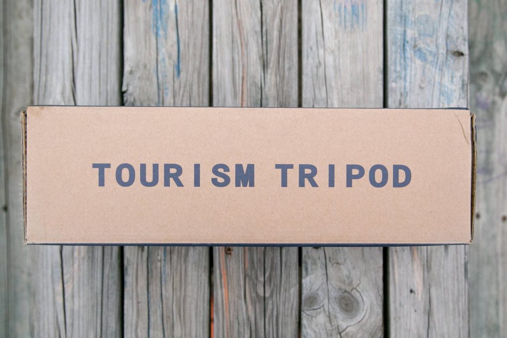 A cardboard box labeled TOURISM TRIPOD sits on a weathered wooden surface.