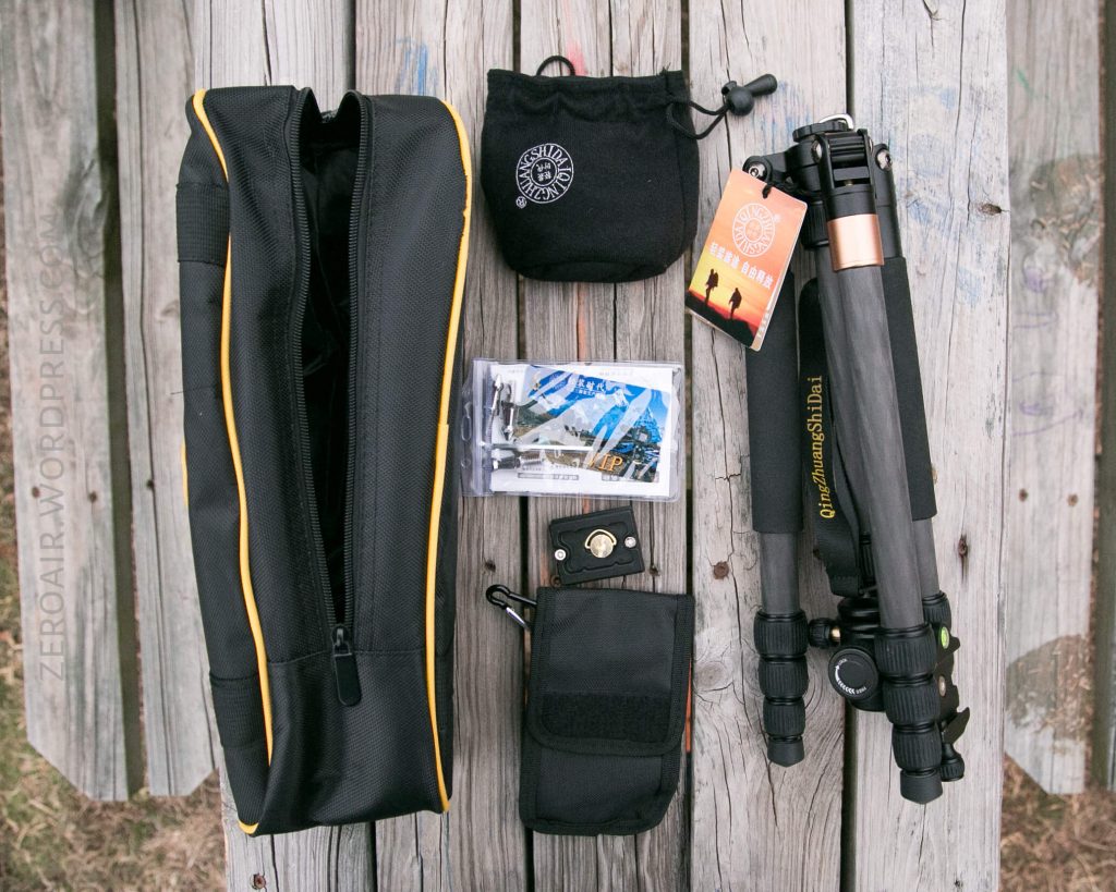 Camera tripod kit items displayed on a wooden surface, including a black carrying case, compact tripod, small pouches, instruction manual, and mounting hardware.