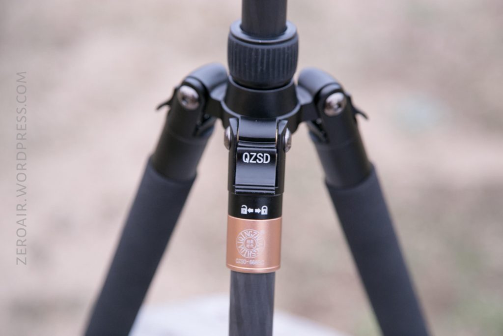 Close-up of a black and bronze tripod leg lock labeled QZSD, showing the locking mechanism and part of the three tripod legs. The background is blurred and neutral in color.