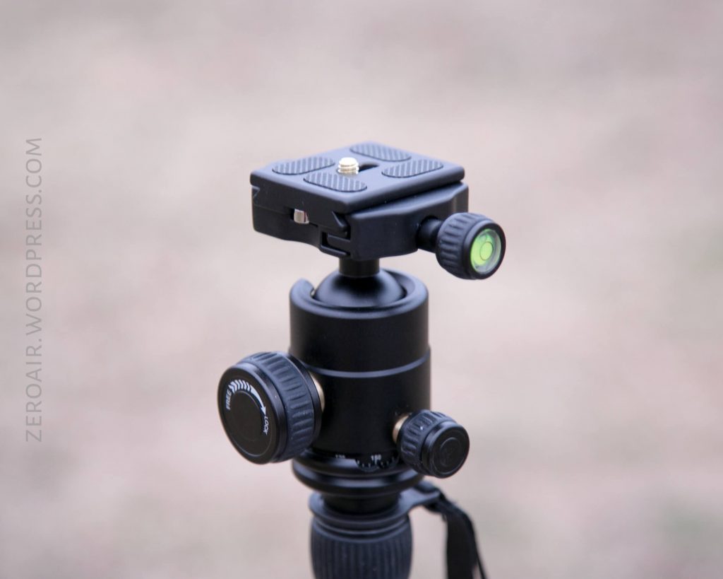 A close-up of a black tripod ball head with adjustment knobs and a bubble level, mounted on a tripod. The background is blurred, and zeroair.wordpress.com is visible on the left.