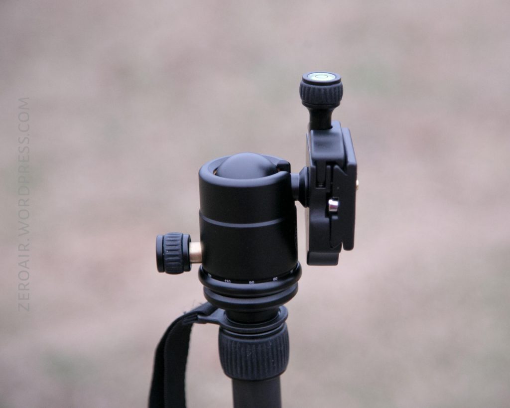 A close-up of a black tripod ball head with adjustment knobs, photographed outdoors on a blurred background. The tripod head is positioned upright, and a strap is visible attached to it.