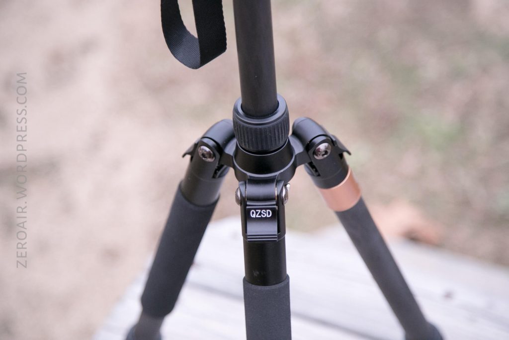 A close-up view of a black QZSD tripod standing on a wooden surface outdoors, showing the tripods central joint, foam leg covers, and part of a tan strap. The background is blurred.