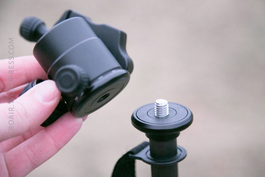 A hand holds a black camera mount above a tripod with a visible screw on top, ready to attach the mount. The background is out of focus.
