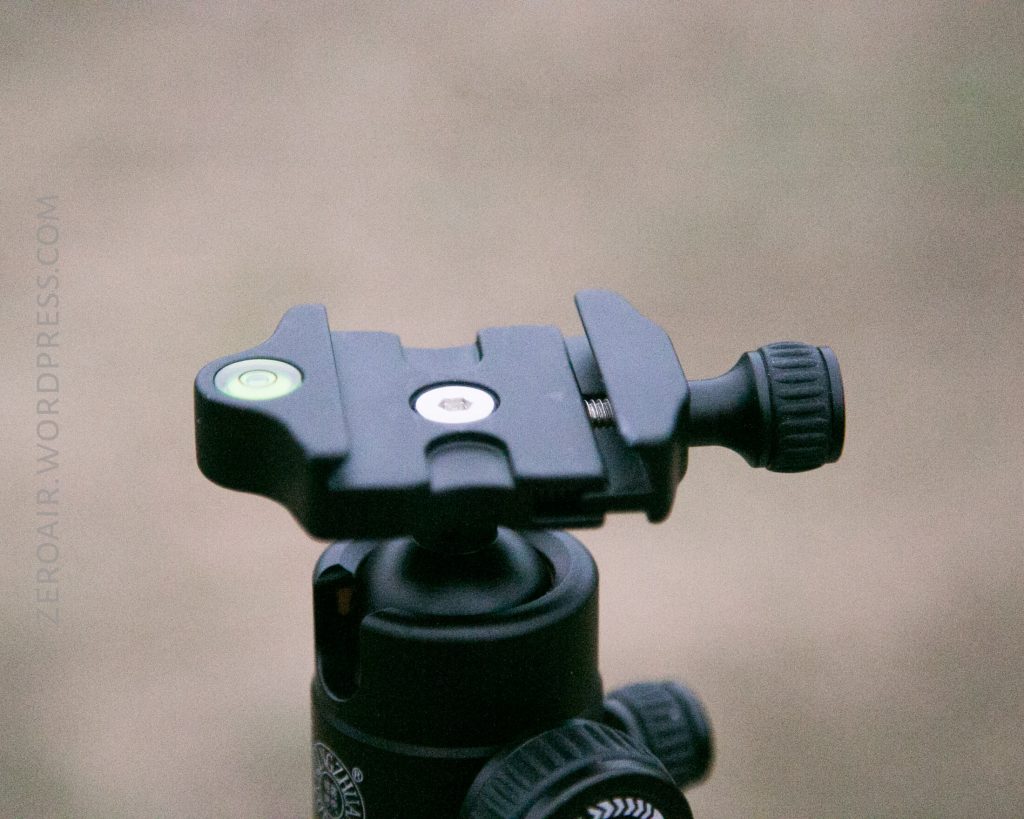 A close-up view of a tripod quick release plate with a built-in bubble level and a tightening knob, mounted on top of a ball head. The background is out of focus.