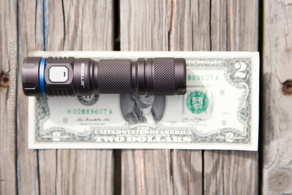 A compact black flashlight is placed horizontally on top of a two-dollar bill, both resting on a wooden surface with visible grain and knots. The flashlight covers part of the bill.