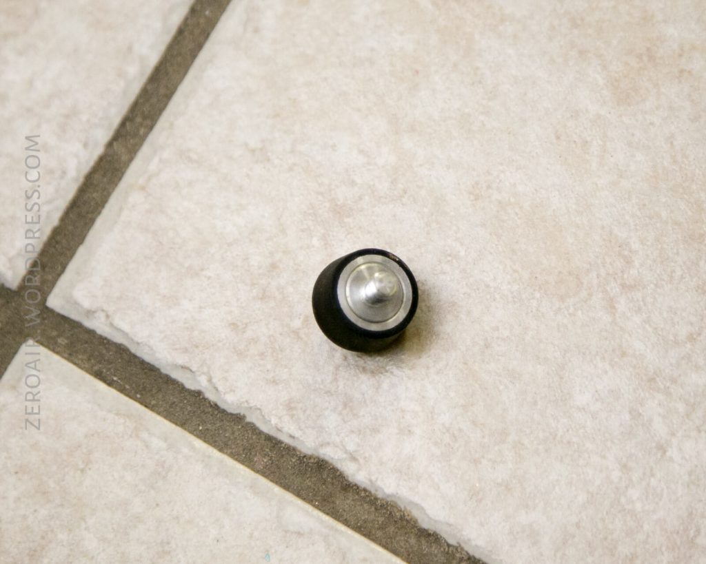 A single wireless earbud with a metallic tip and black casing lies on a tiled floor with beige and gray grout lines.