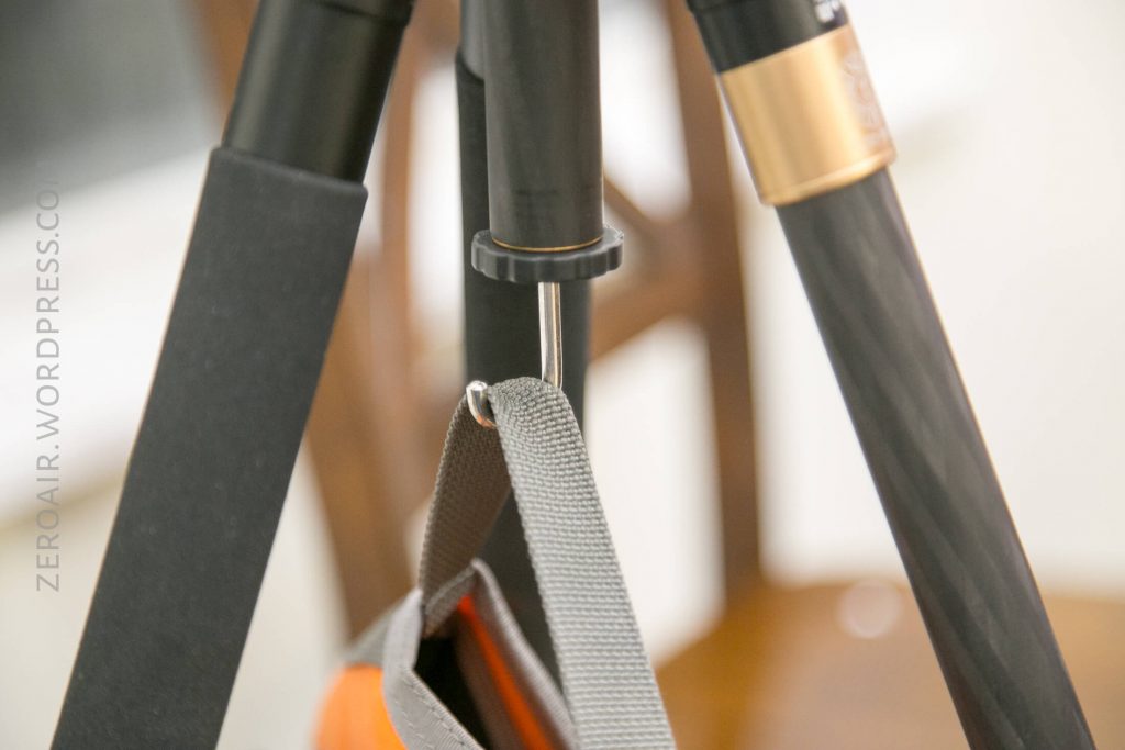 A close-up of a tripod leg with a hook, holding a gray strap from a weighted bag. The background is out of focus, and zeroair.wordpress.com is visible on the left side of the image.
