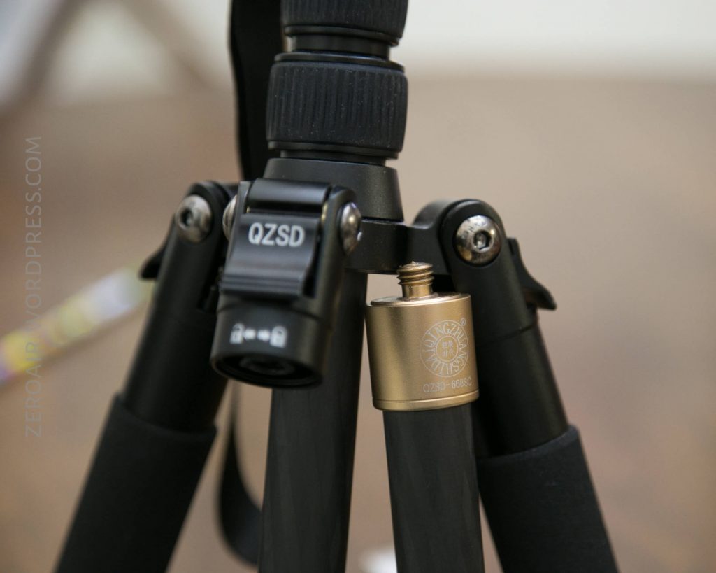 Close-up of a black tripod with a gold-colored metal object, possibly an accessory or small container, attached to one of the legs. The brand QZSD is visible on the tripod head.