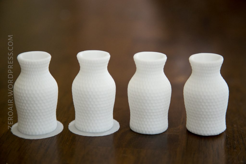 Four small, white, 3D-printed vases with a textured, honeycomb-like surface are arranged in a row on a brown wooden surface. The leftmost vase has a circular base plate attached.