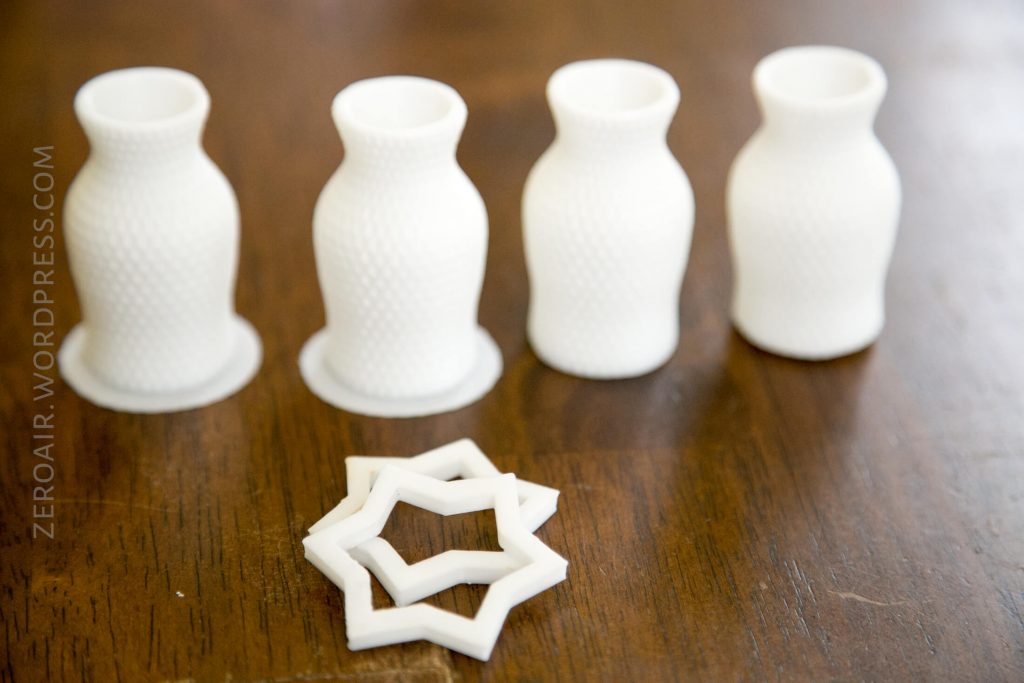 Four small, white, textured 3D-printed vases are lined up on a wooden surface, with two interlocking, star-shaped 3D-printed objects placed in front of them.