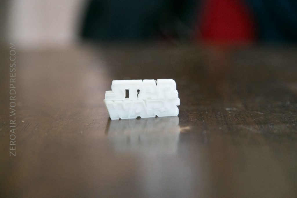 A small white 3D-printed object spelling out STAR WARS in block letters sits on a wooden surface. The background is out of focus, and text on the left reads ZEROAIR.WORDPRESS.COM.
