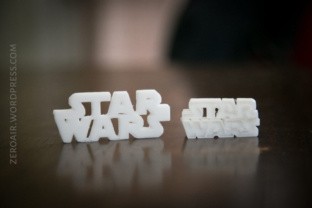Two white 3D-printed objects display the Star Wars logo in block letters, one larger and one smaller, placed on a reflective brown surface. The background is blurred.
