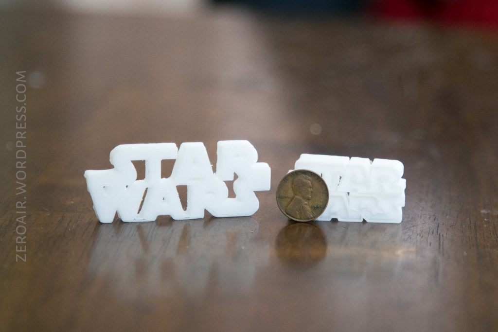 Two white 3D-printed objects shaped like the Star Wars logo are placed on a wooden surface with a coin standing upright between them for scale. The background is out of focus.
