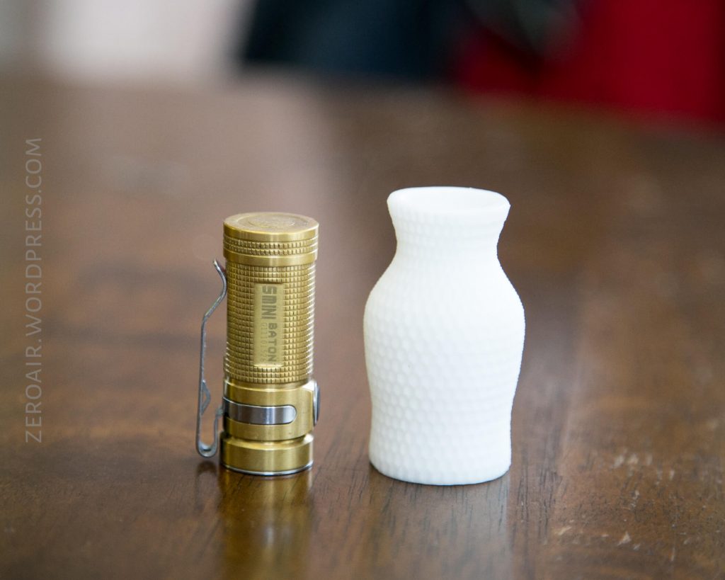 A small brass flashlight with a pocket clip stands upright next to a white, textured, 3D-printed vase on a wooden surface. Both objects are in focus, with a blurred background.