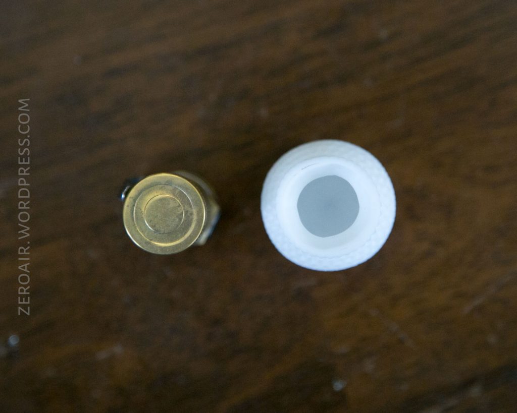 A brass AA battery is shown next to a white, hollow golf ball on a wooden surface, viewed from above. The battery is on the left and the golf ball is on the right. ZEROAIR.WORDPRESS.COM is visible on the left edge.