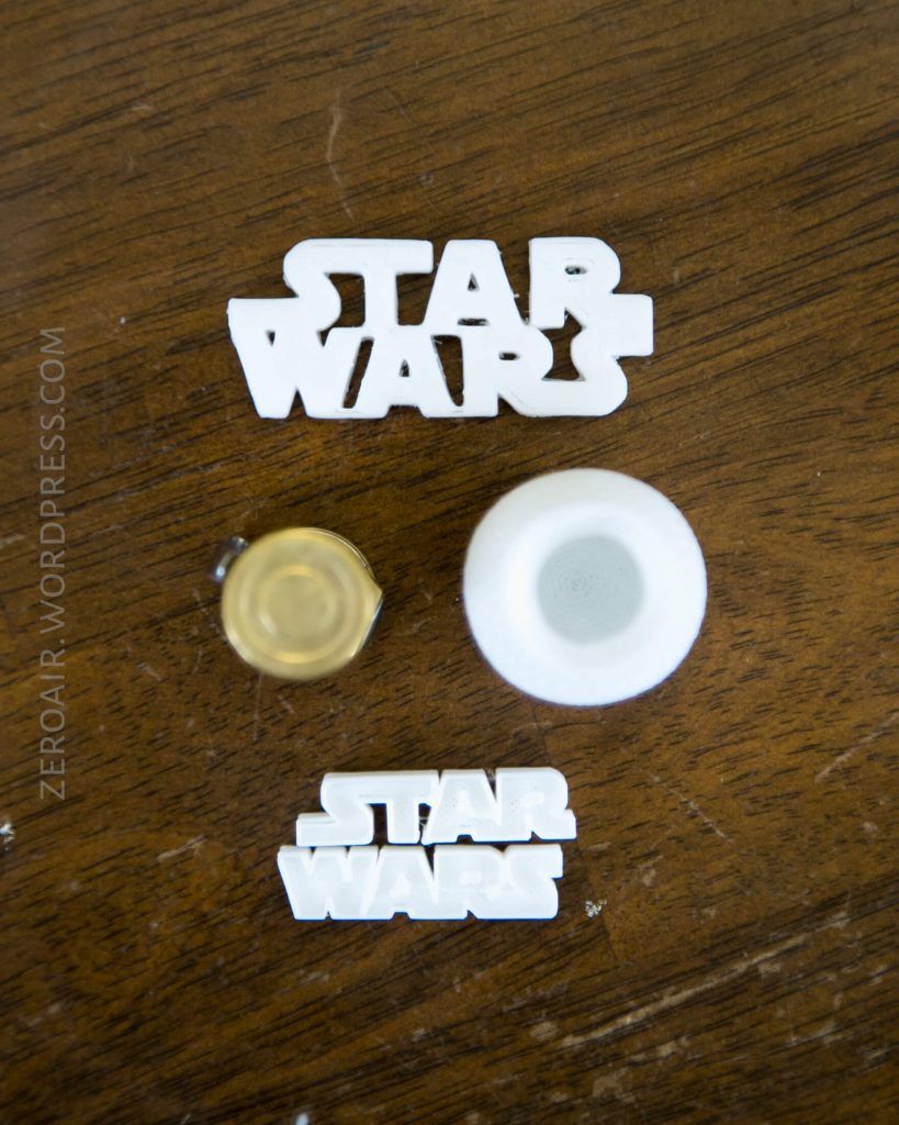 Two white 3D-printed Star Wars logos, a small round container with a clear top, and another white lid are placed on a wooden surface. ZERODAIR.WORDPRESS.COM is visible along the left side.