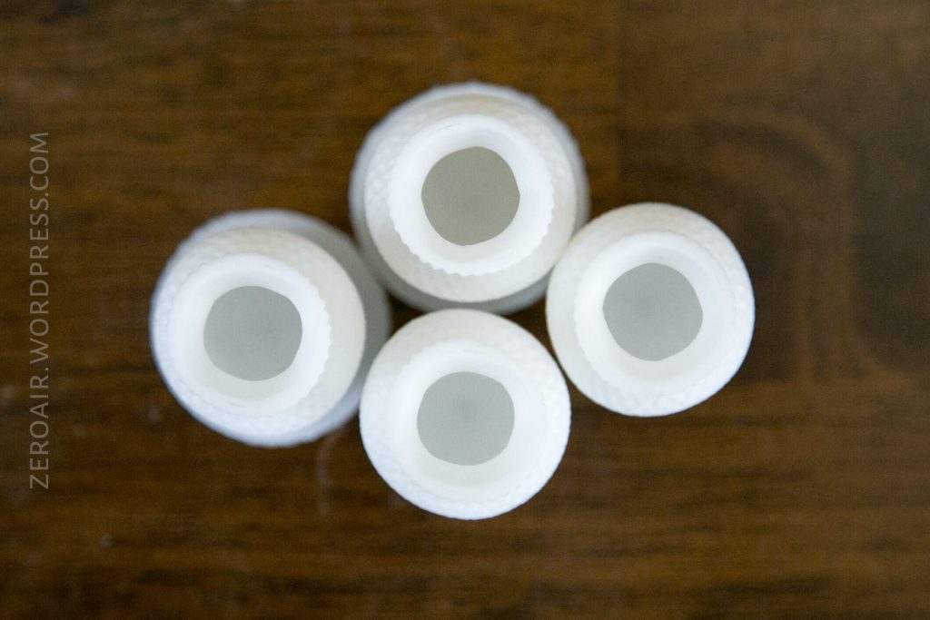 Four white plastic bottle openings viewed from above, arranged in a close group on a wooden surface. The bottles have textured sides, and the image has a watermark on the left side that reads ZEROAIR.WORDPRESS.COM.